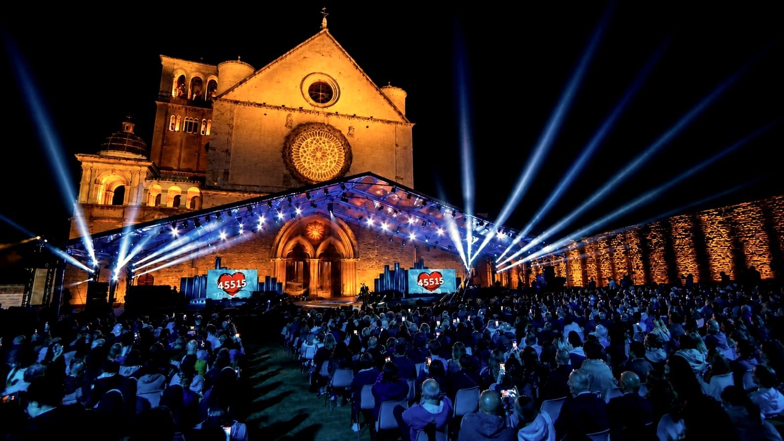 La facciata della Basilica di San Francesco ad Assisi illuminata di notte, con fasci di luce nel cielo.